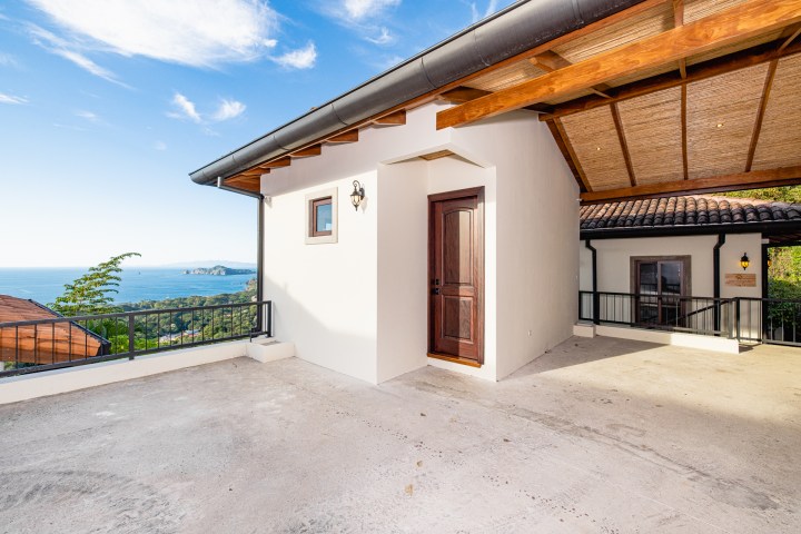 House entrance with wooden door, covered patio, ocean view, and clear blue sky.