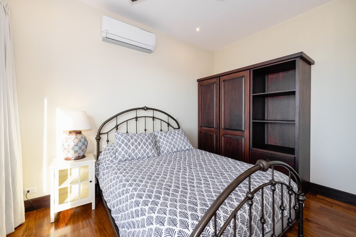 Bedroom with metal bedframe, patterned bedding, dark wood cabinet, side table with lamp, and wall-mounted AC.