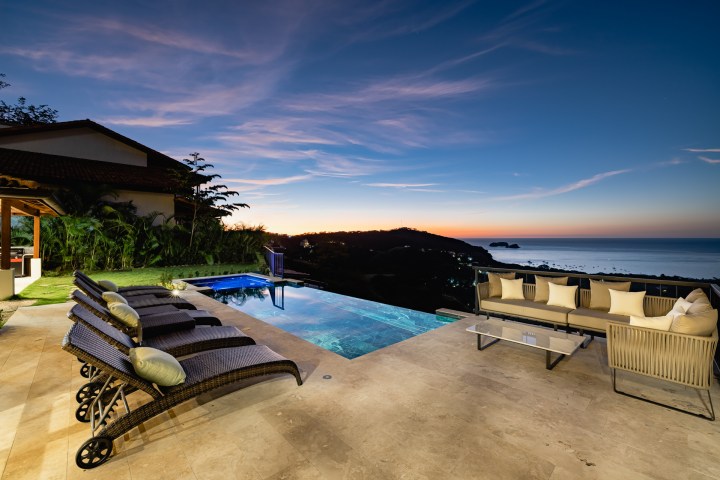 Sunset view from patio with infinity pool, lounge chairs, and ocean in the background.