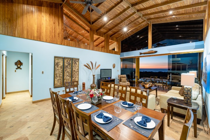 Spacious living area with wooden ceiling, dining table set, and ocean view at sunset.