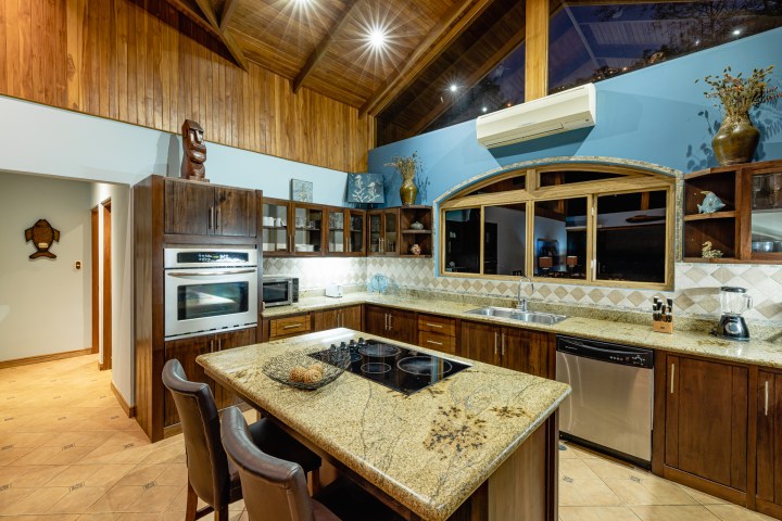 Wooden kitchen with island, oven, and blue wall. Granite countertops, modern appliances, and high ceiling.