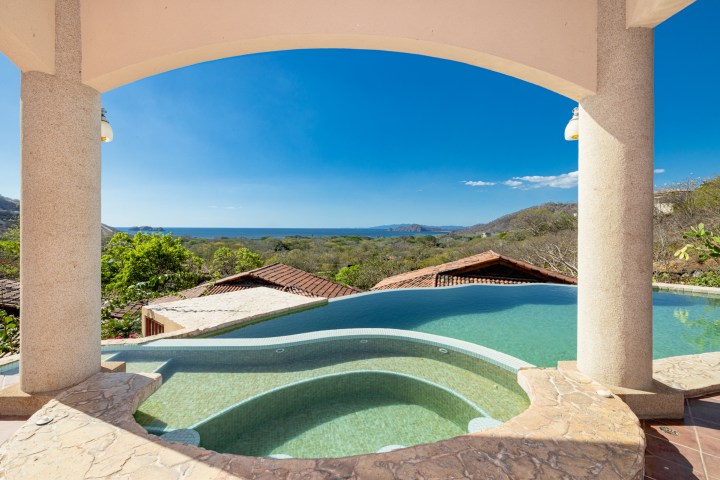 Infinity pool with ocean view, framed by stone pillars and terracotta roofs under a clear blue sky.