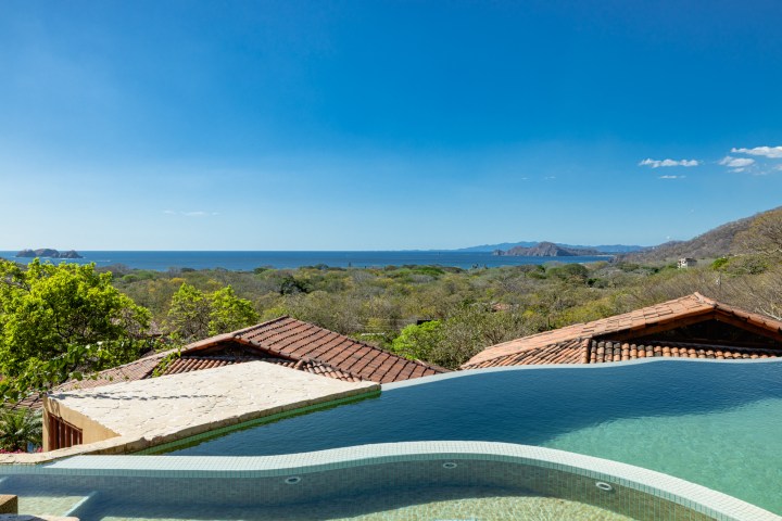Infinity pool overlooking trees and ocean with mountains in the background under a clear blue sky.