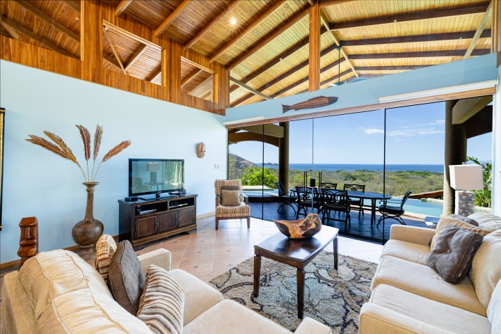 Spacious living room with ocean view, beige sofas, wooden ceiling, and glass doors opening to a terrace.
