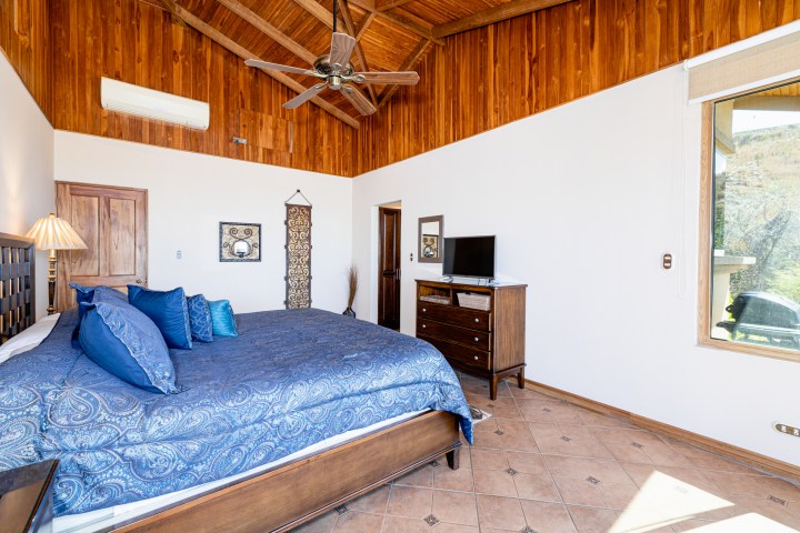 Bedroom with a large bed, blue bedding, wooden ceiling, TV on dresser, and a large window.