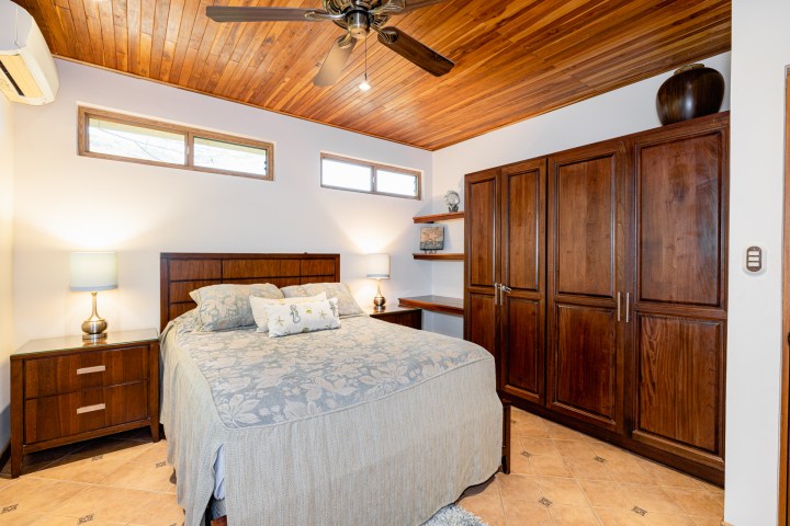 Bedroom with wooden ceiling, fan, bed, two lamps, and wooden wardrobe.