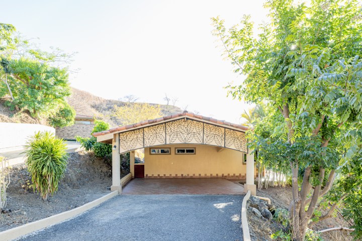 A small beige house with a decorative arch, surrounded by trees and greenery.