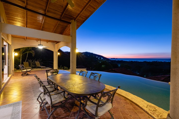 Covered patio with dining set overlooking infinity pool and scenic sunset view.