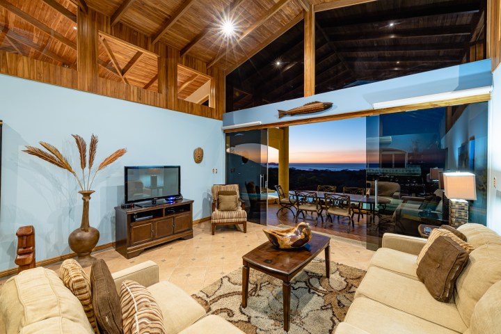 Spacious living room with wooden ceiling, large TV, beige sofas, and glass doors leading to a sunset view.