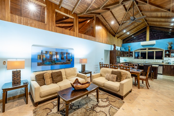 Spacious living and dining area with wooden ceiling, sofas, dining table, and kitchen in the background.