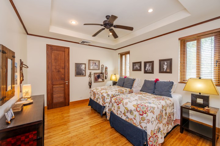 Bedroom with twin beds, floral bedding, wooden floor, and ceiling fan, decorated with framed photos and lamps.