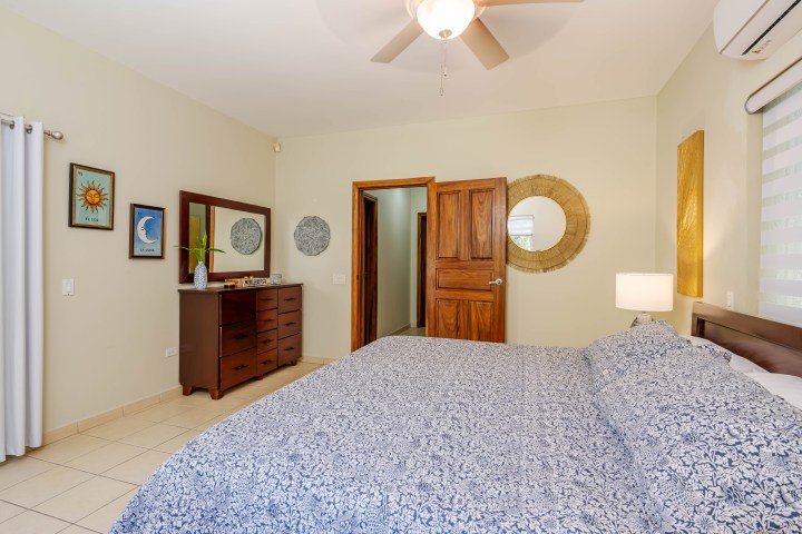 Bedroom with blue patterned bedspread, wooden dresser, large mirror, ceiling fan, and framed artwork on walls.