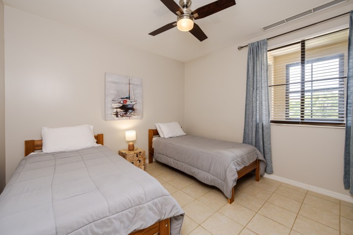 Simple bedroom with two single beds, a ceiling fan, and a window with blue curtains.