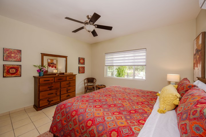 Cozy bedroom with colorful bedspread, ceiling fan, wooden dresser, and window with blinds.