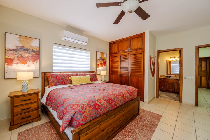 Bedroom with patterned bedspread, wooden furniture, artwork, ceiling fan, and open door to bathroom.
