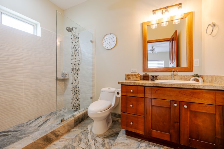 Modern bathroom with wood vanity, large mirror, glass shower, and toilet on a marble floor.