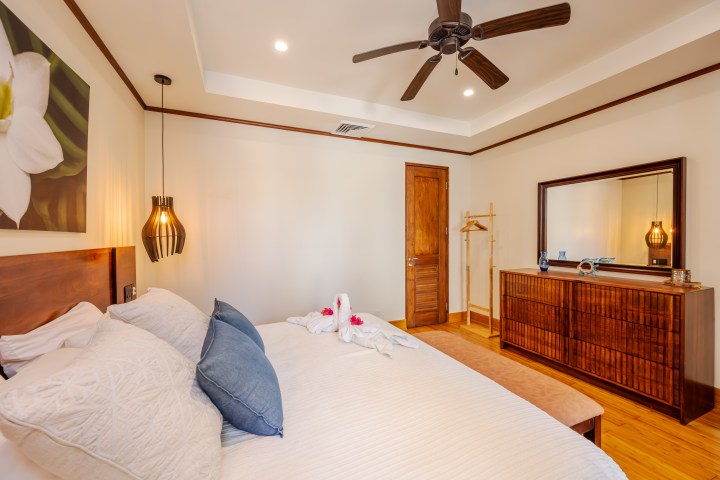Cozy bedroom with wooden furniture, bed with cushions, ceiling fan, and decorative swan towels.