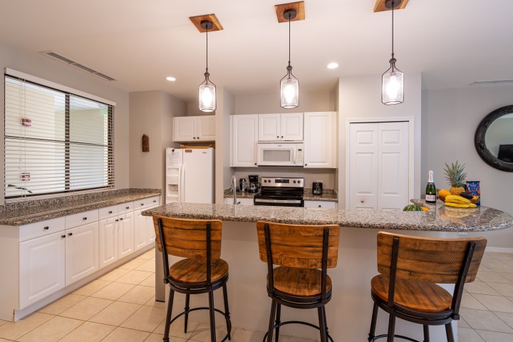 Modern kitchen with three wooden bar stools, granite countertop, hanging lights, and white cabinets.