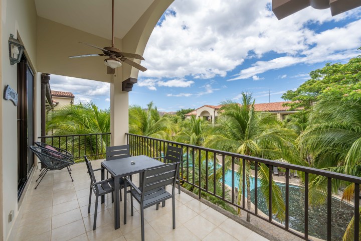 Balcony with table, chairs, ceiling fan, view of palm trees and pool under a cloudy sky.