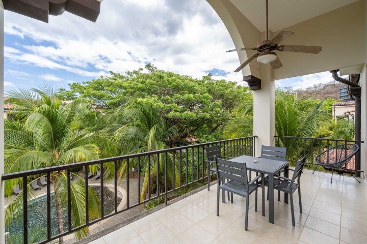 Balcony with table, chairs, ceiling fan, overlooking lush trees and cloudy sky.