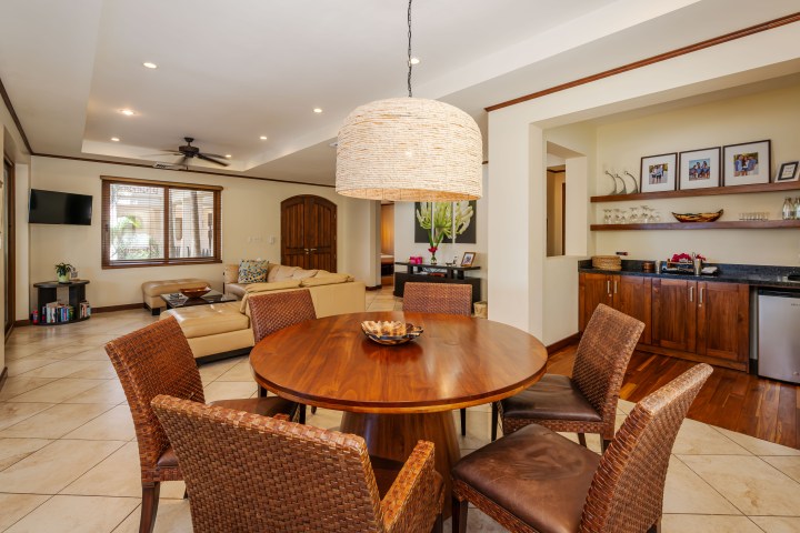 Dining area with round wooden table, wicker chairs, and a large hanging lamp in an open living room.