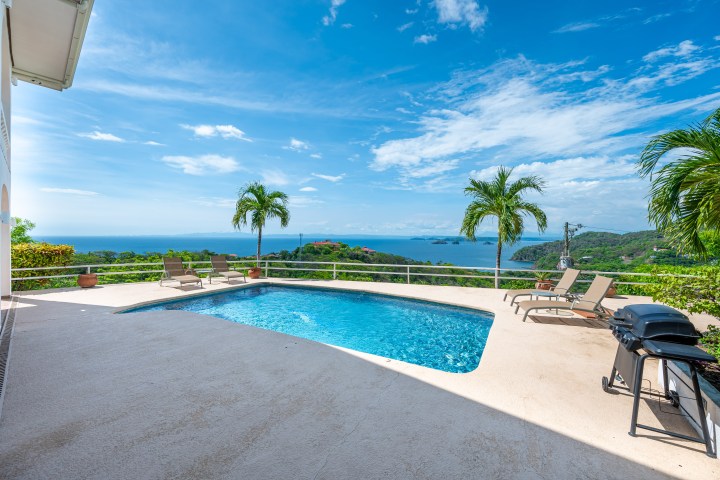 Poolside view with loungers, palm trees, and ocean in the background under a blue sky.