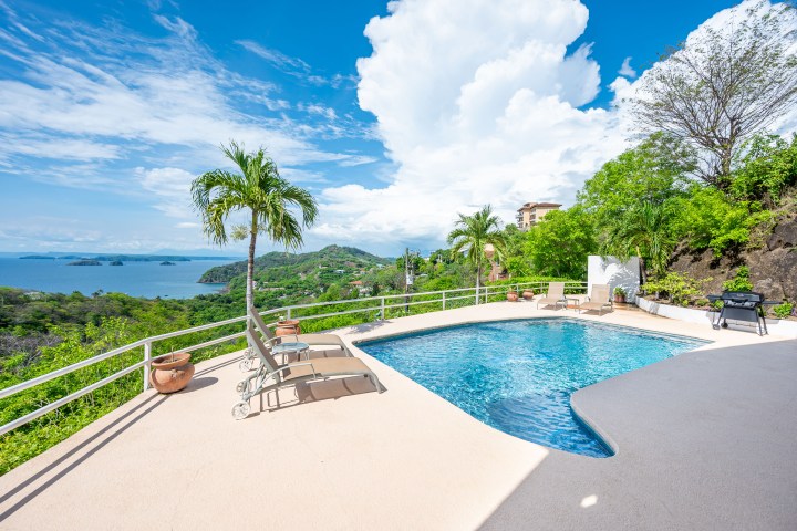 Poolside view with lounge chairs, overlooking a tropical landscape and ocean under a blue sky.