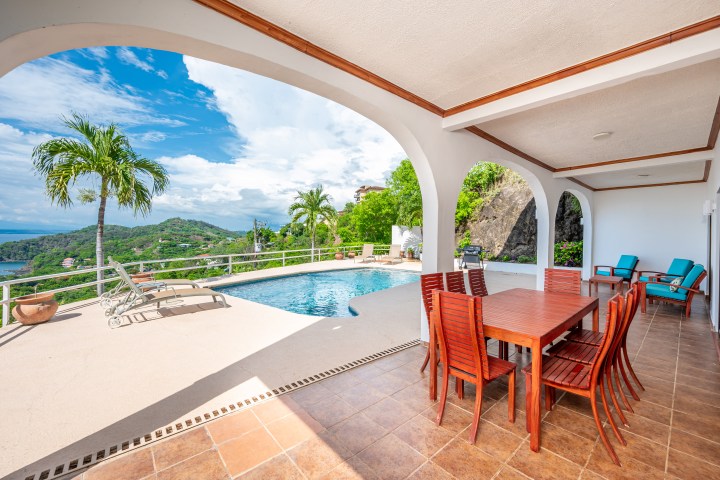 Covered patio with wooden dining set overlooking pool and tropical landscape.
