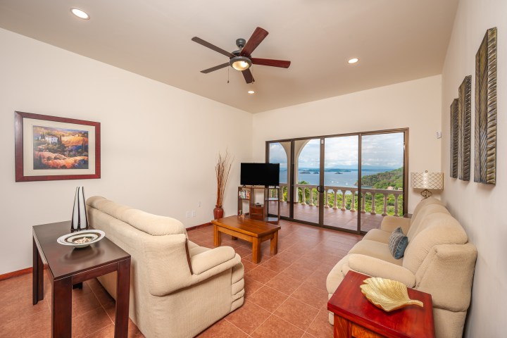 Spacious living room with beige sofas, a ceiling fan, and large windows overlooking a scenic ocean view.