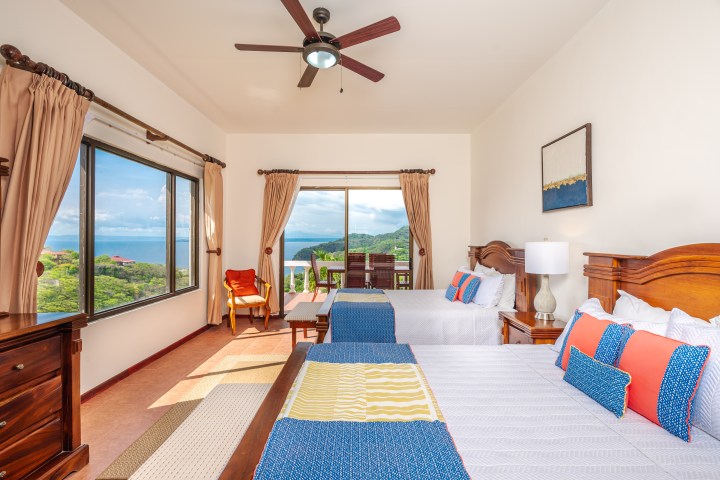 Bright bedroom with two beds, ocean view from large windows, fan, and wooden furniture.