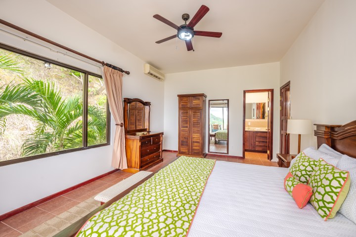 Bright bedroom with wooden furniture, large window view of greenery, and ceiling fan.