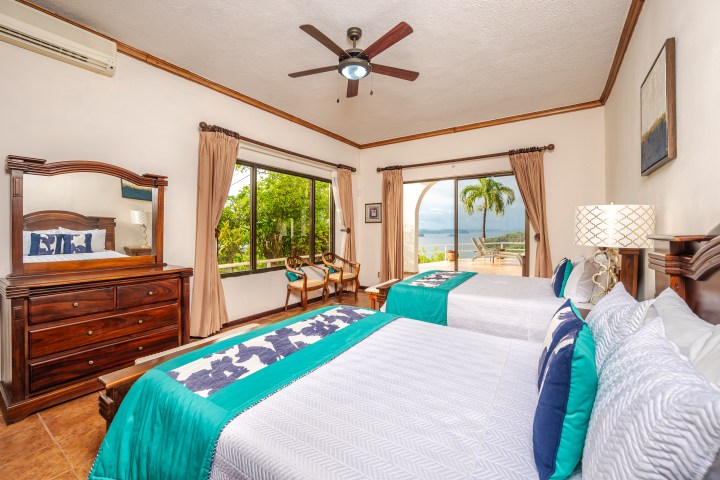 Bright bedroom with two beds, wooden furniture, and ocean view through large window and sliding door.