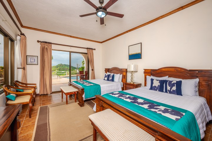 Hotel room with two beds, wooden furniture, ceiling fan, and a view of pool and hills through sliding glass door.