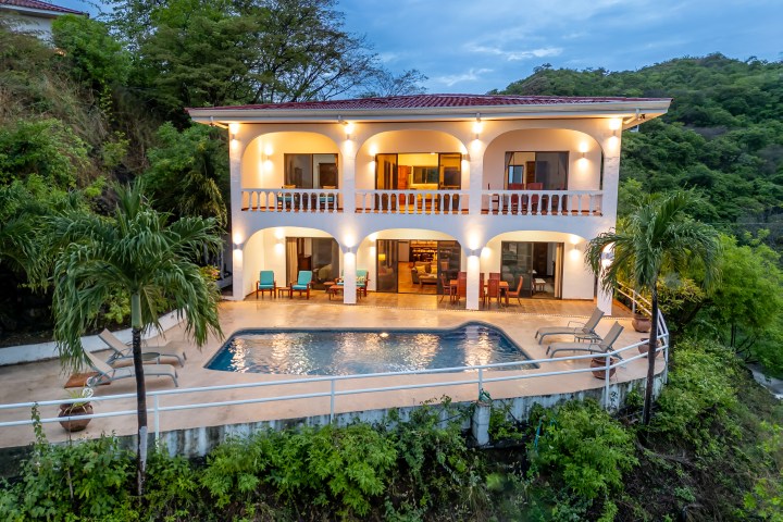Two-story villa with balconies, pool, and lush surroundings at dusk.