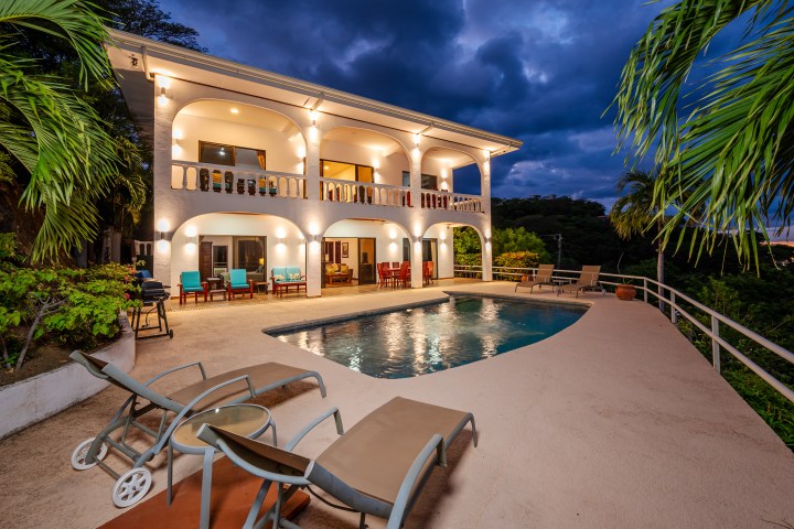 Two-story villa with arches, lit at dusk, overlooking a pool and surrounded by palm trees.