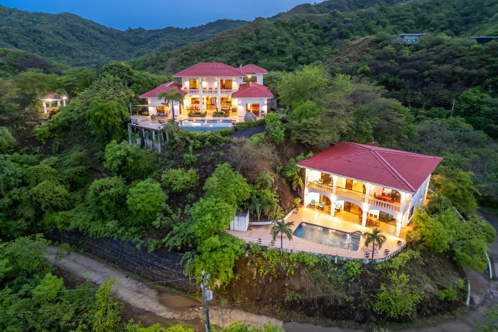 Aerial view of two illuminated houses with red roofs on a forested hillside at dusk.