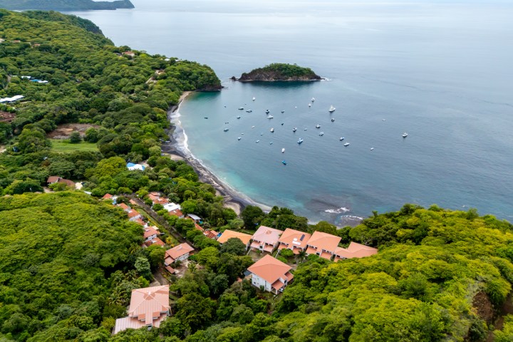 Aerial view of houses among lush greenery near a bay with anchored boats and small island.
