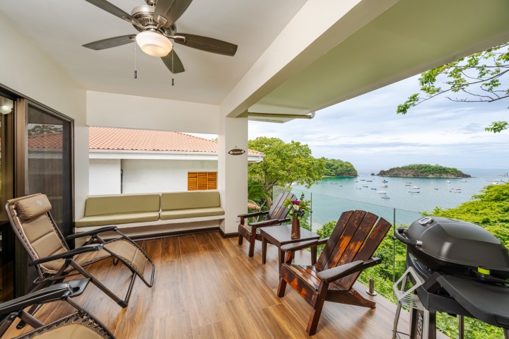 Balcony with seating, ceiling fan, and grill overlooking a bay with boats and lush greenery.
