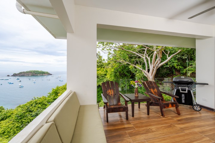 Balcony view with two chairs, table, and grill overlooking ocean and island.