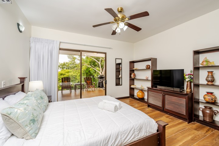 Bedroom with wooden floors, ceiling fan, TV, and sliding doors opening to a balcony with chairs and greenery.