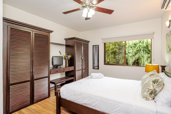 Bedroom with wooden furniture, white bedding, a ceiling fan, and a window with greenery outside.