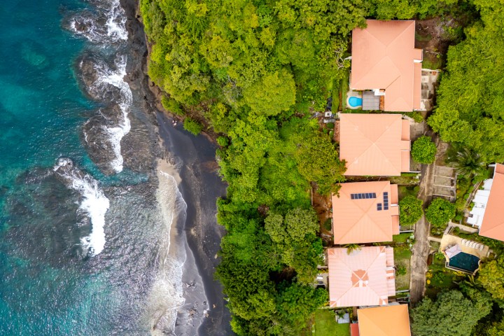 Aerial view of coastal houses with red roofs by a blue ocean and dense green forest.