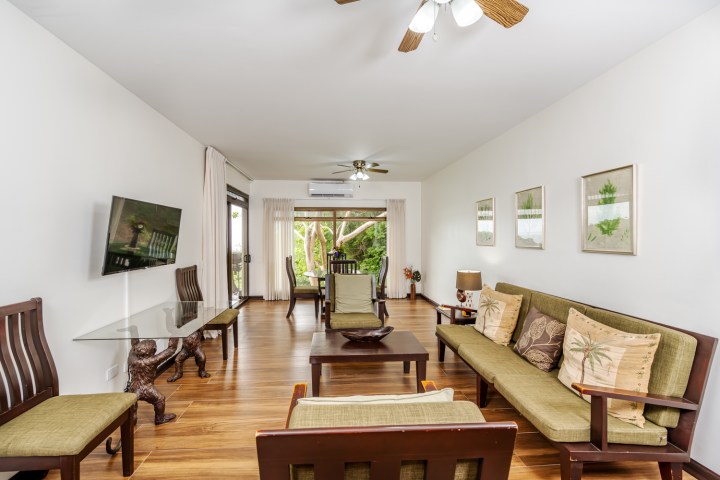Bright living room with wooden floors, green sofas, wooden furniture, ceiling fan, and wall art.