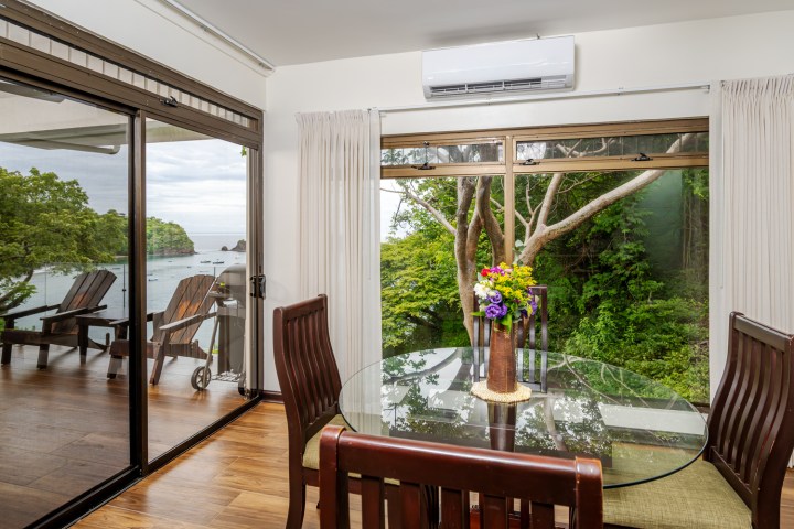 Dining area with glass table, forest view, and balcony overlooking a bay with chairs.
