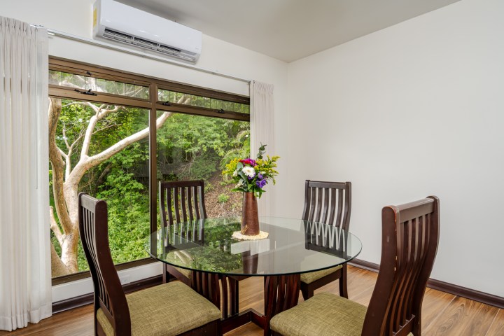 Dining area with round glass table, four chairs, and window view of trees.