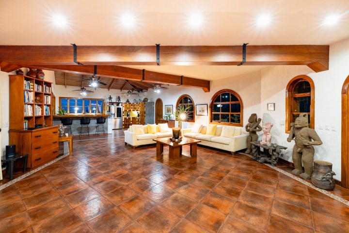 Spacious living room with wooden beams, white sofas, statues, and a kitchen in the background.