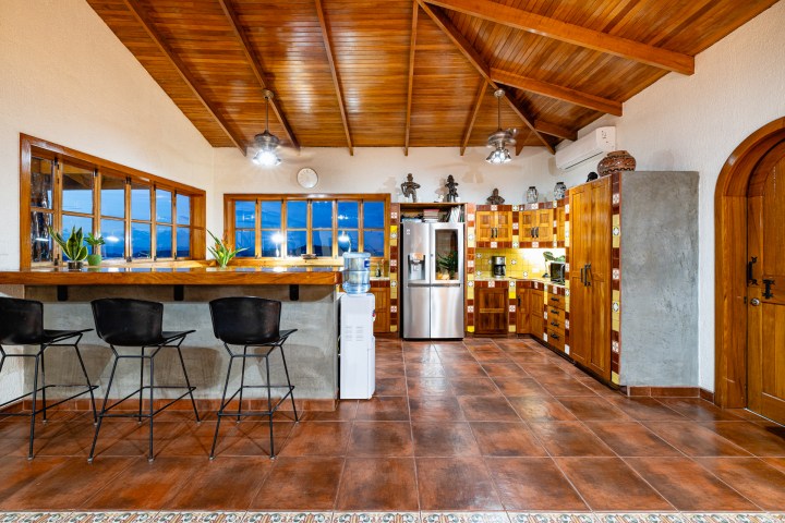 Rustic kitchen with wooden cabinets, bar stools, fridge, and tiled floor and ceiling.