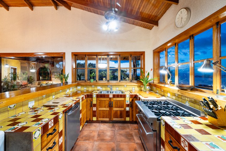 Rustic kitchen with wooden cabinets, colorful tiles, and large window showing a scenic view.