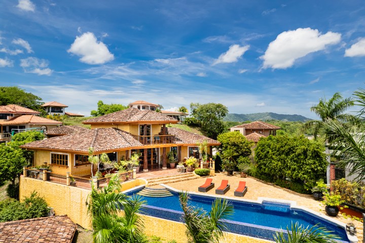 Aerial view of a luxury house with pool, surrounded by greenery and under a clear blue sky.