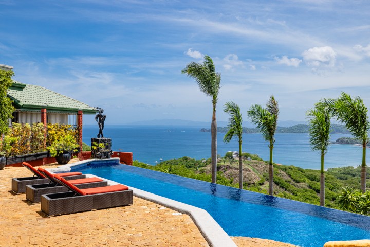 Infinity pool with ocean view, palm trees, lounge chairs, and a sculpture on a sunny day.